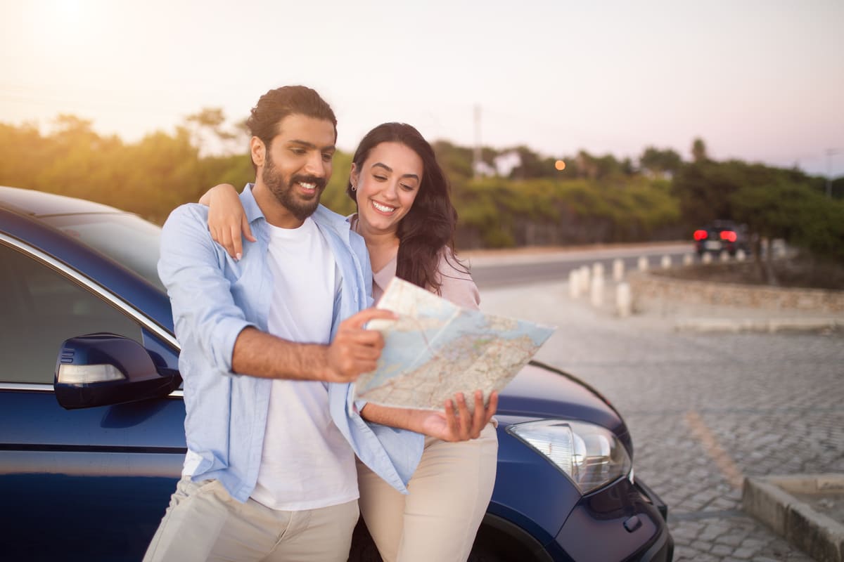 Casal sorridente em uma estrada ao pôr do sol, consultando um mapa ao lado de um carro, planejando a viagem para um dos seus lugares para passar a lua de mel dos sonhos.