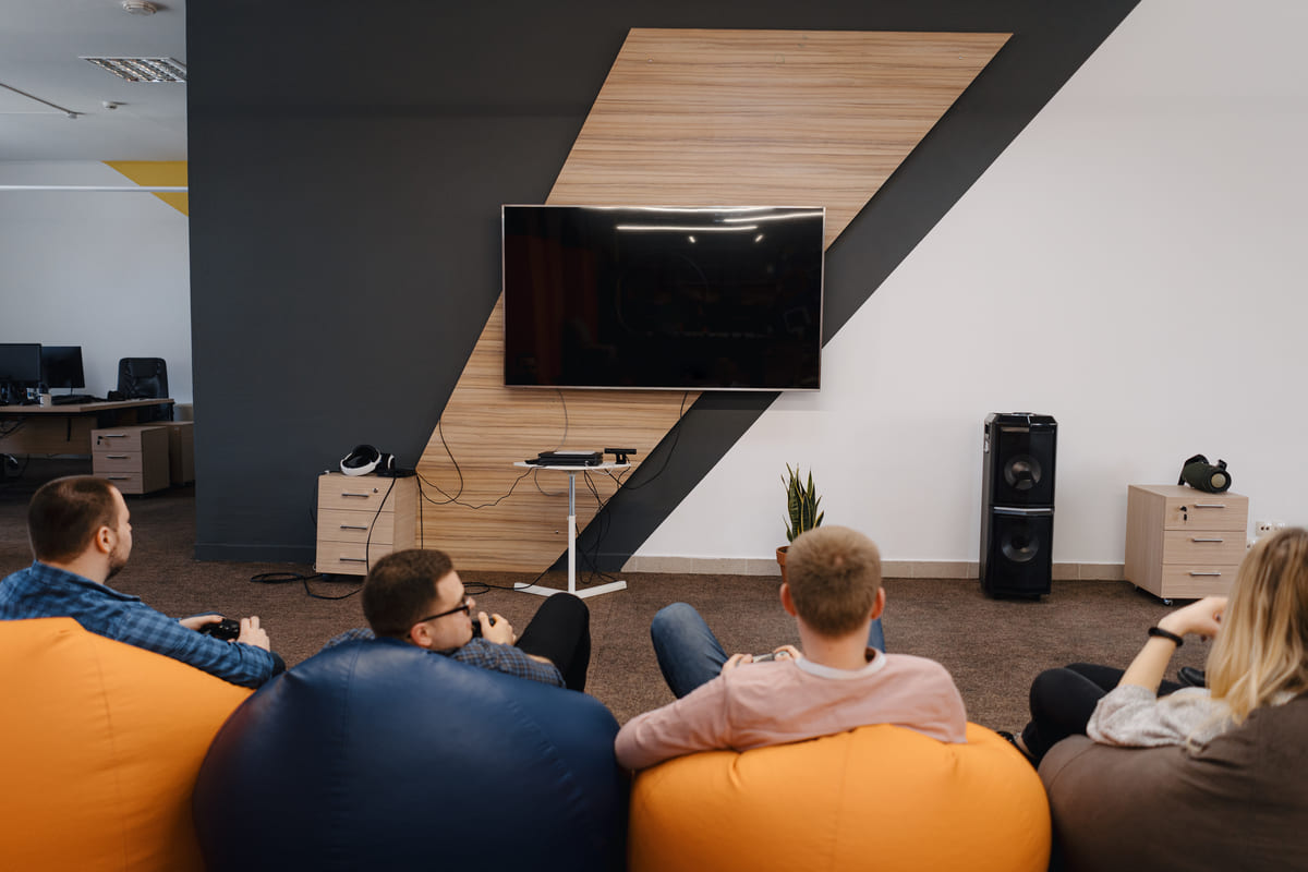 Amigos sentados em pufes coloridos jogam videogame em uma sala de tv com painel de madeira moderno.