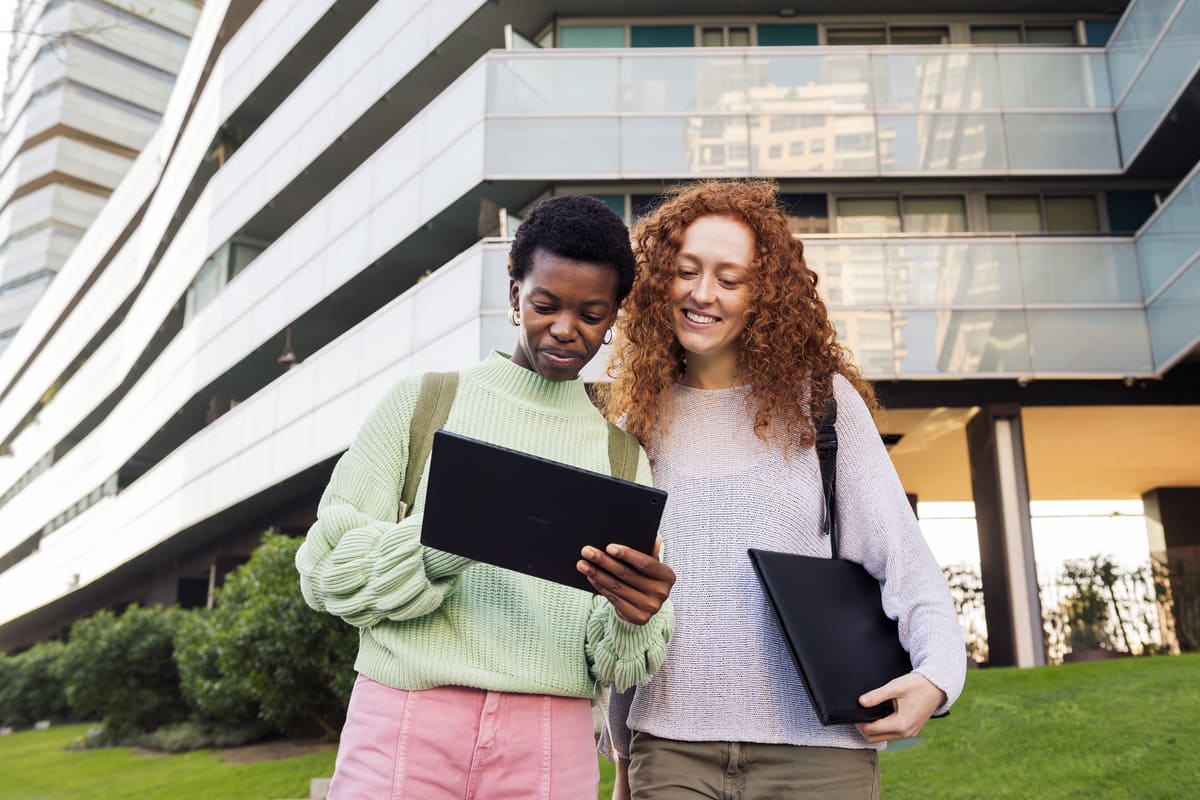 Duas jovens sorridentes olhando juntas para um tablet em frente a um prédio moderno, ilustrando o planejamento do futuro e a busca por oportunidades com um consórcio para estudos.
