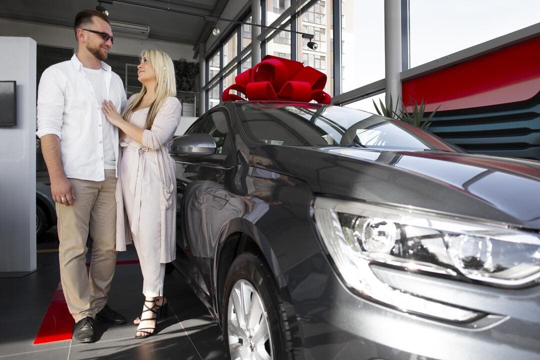 Carro novo ou usado: uma mulher e um homem comprando um carro novo na cor cinza, com um laço vermelho em cima.