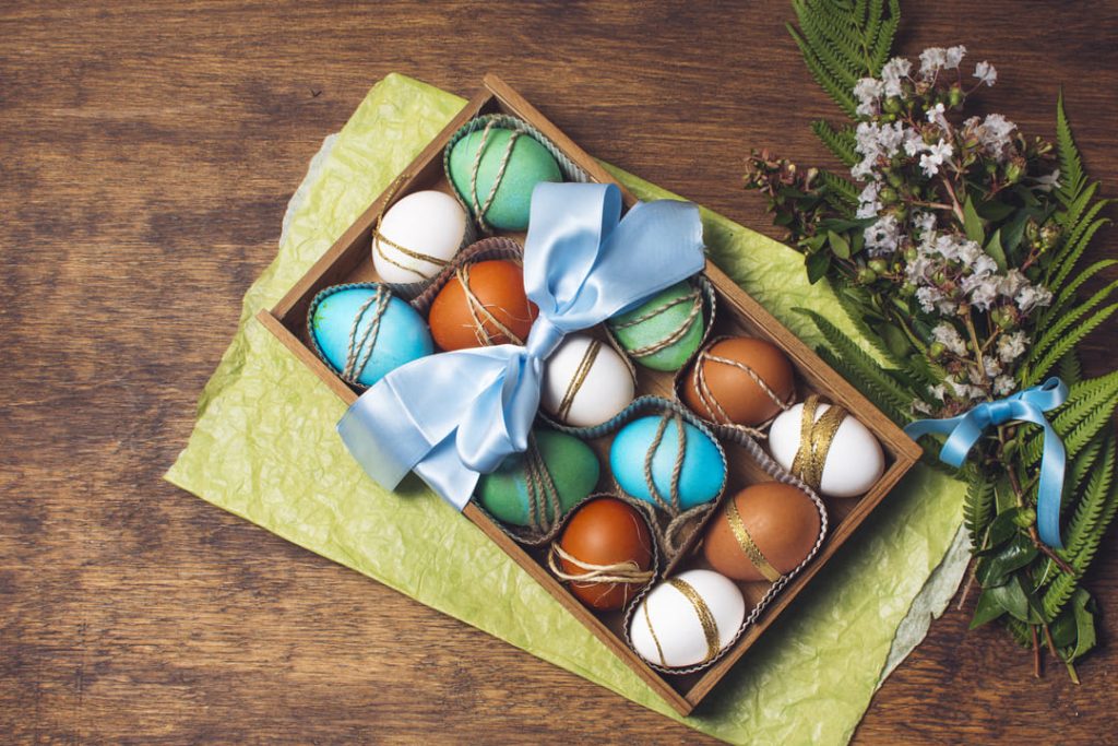 Páscoa: uma caixa com vários ovos decorados e com uma fita azul clara amarrada e umas flores lilás ao lado.