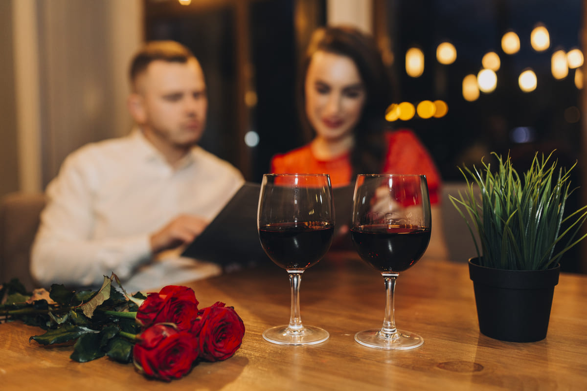 Duas taças de vinho tinto e um buquê de rosas vermelhas sobre a mesa, com um casal ao fundo em um jantar romântico, ilustrando experiências como dicas de presente para o Dia dos Namorados.