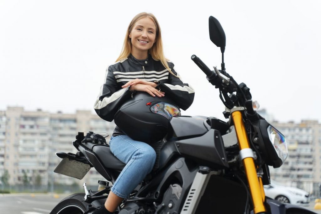 Moto para comprar: uma mulher loira está sentada em uma moto preta, segurando o capacete nas mãos e sorrindo.
