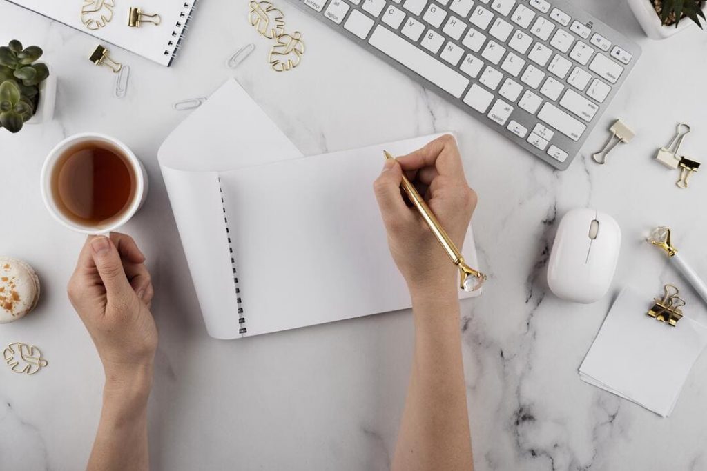Dicas financeiras organize as finanças para o próximo ano: uma pessoa com um caderno mais uma caneta dourada na mão e um teclado branco e cinza do lado e uma xícara de café. 