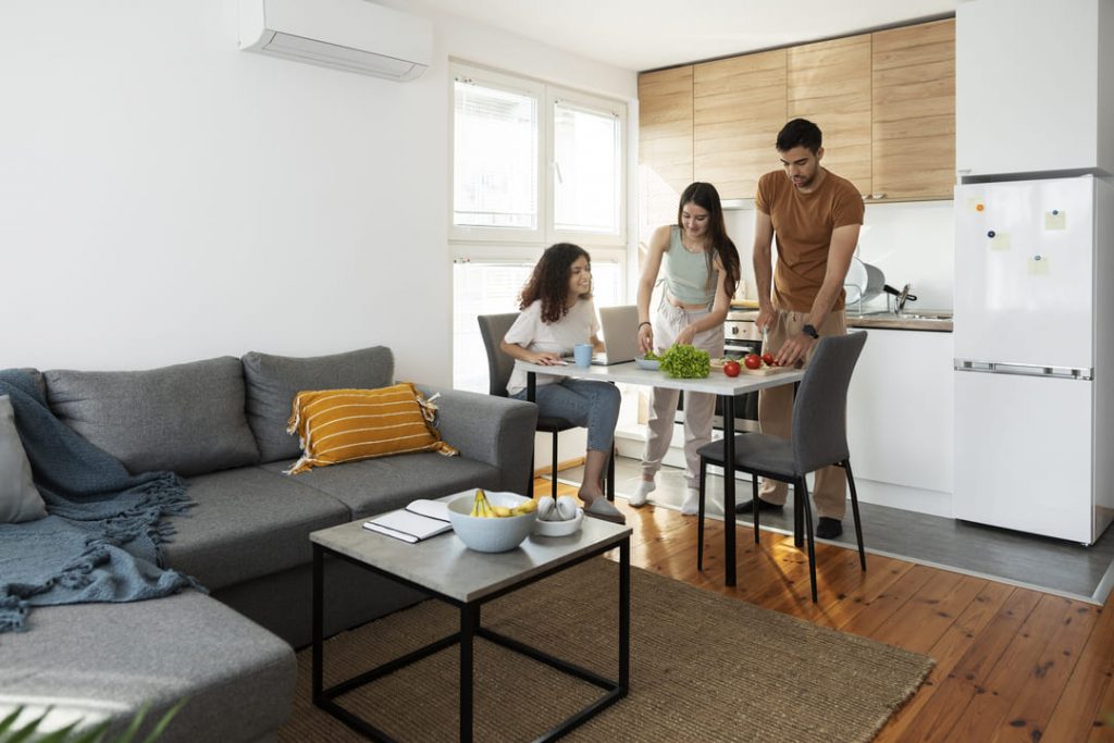Comprar apartamento estúdio: três pessoas conversando, uma sentada na cadeira em frente a uma mesa e duas em pé ao lado. Tem um sofá cinza e uma mesa de centro branca.