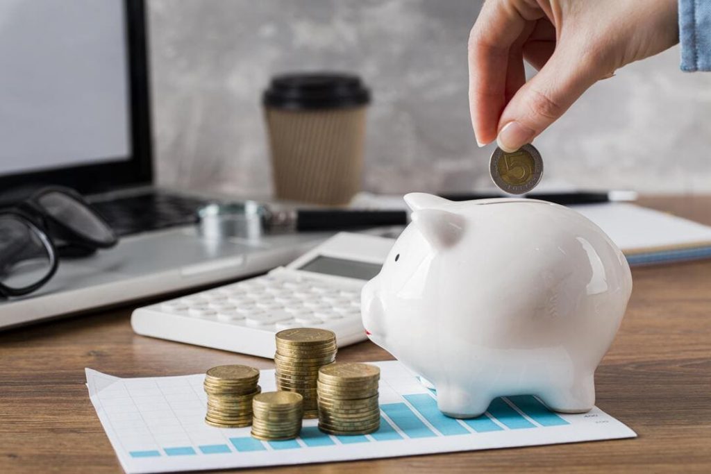 O que é fundo comum no consórcio: uma pessoa colocando dinheiro em um cofrinho de porquinho rosa e ao lado várias moedas douradas e uma calculadora.