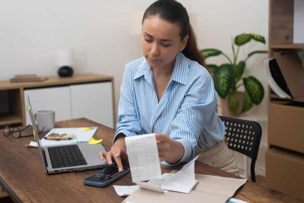 Método 50 30 20: uma mulher de pele clara, segurando uma conta em uma mão e fazendo contas com o celular na outra, com um notebook ao lado.