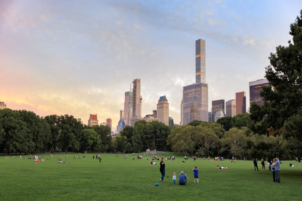 A imagem mostra uma vista do Central Park em Nova York ao entardecer. No primeiro plano, há uma grande área gramada onde várias pessoas estão sentadas, caminhando ou brincando, aproveitando o ambiente do parque. Algumas crianças estão jogando bola enquanto outras pessoas estão relaxando ou interagindo umas com as outras. Ao fundo, destaca-se o horizonte de Manhattan com seus arranha-céus icônicos. Os edifícios são altos e imponentes, com design moderno e clássico misturados. O céu está com um tom suave de fim de tarde, com nuances de laranja e azul, criando um ambiente calmo e agradável. A vegetação ao redor da área gramada é densa, com muitas árvores que adicionam um toque de verde à paisagem urbana.