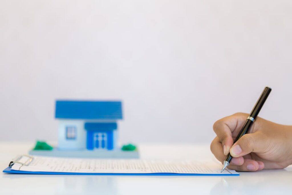 A imagem mostra uma pessoa assinando um documento sobre uma mesa branca. O documento está preso a uma prancheta azul. Ao fundo, há uma pequena maquete de uma casa com telhado azul, representando simbolicamente o setor imobiliário. A mão da pessoa segura uma caneta preta enquanto assina o papel, sugerindo que se trata de um contrato ou documentação relacionada a imóveis. O fundo desfocado destaca a ação de assinatura como o foco principal da imagem.