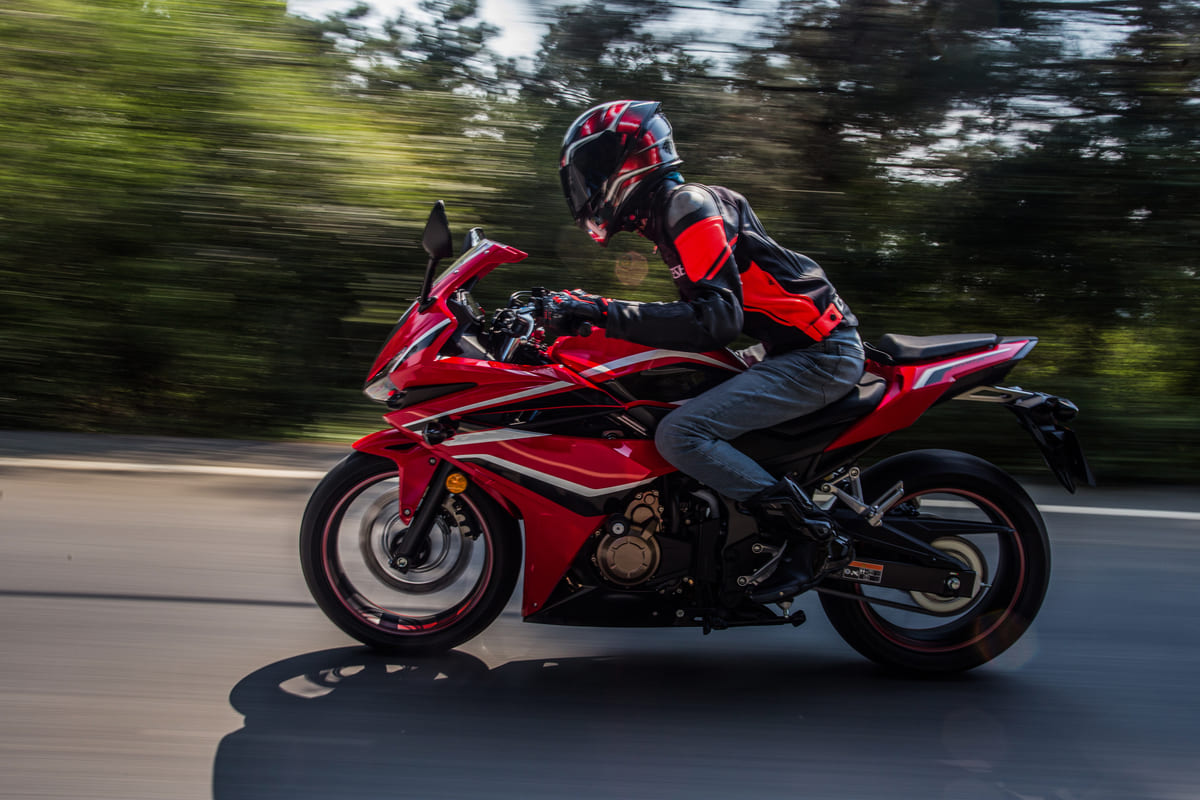 Homem pilotando moto esportiva na cor vermelha em uma estrada pavimentada com árvores ao fundo, em alusão às motos em lançamento do ano.
