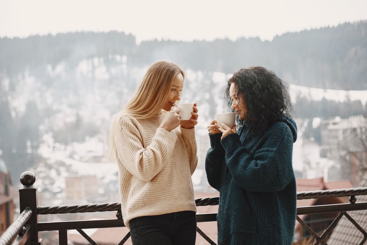 Duas mulheres com suéteres de inverno conversam enquanto tomam uma bebida quente na varanda, com uma paisagem montanhosa ao fundo, desfrutando do clima de um dos charmosos lugares para viajar no inverno no Brasil.