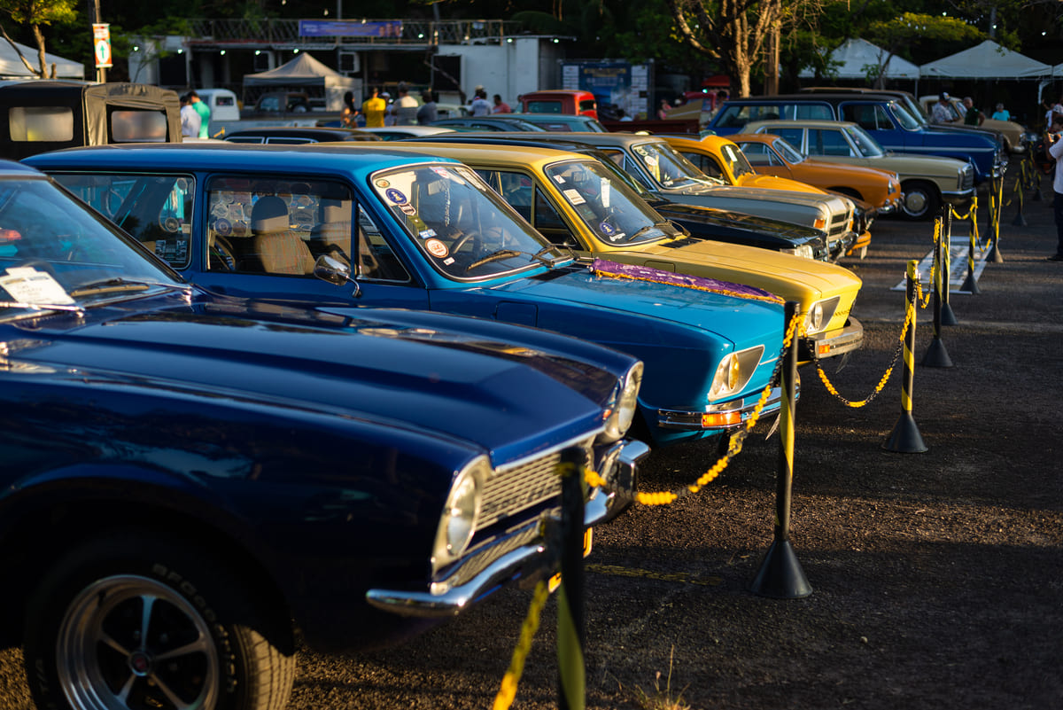 Encontro de carros antigos ao ar livre, com uma fileira de modelos clássicos brasileiros, como Volkswagen Brasília e Variant, estacionados e isolados por uma corda amarela.