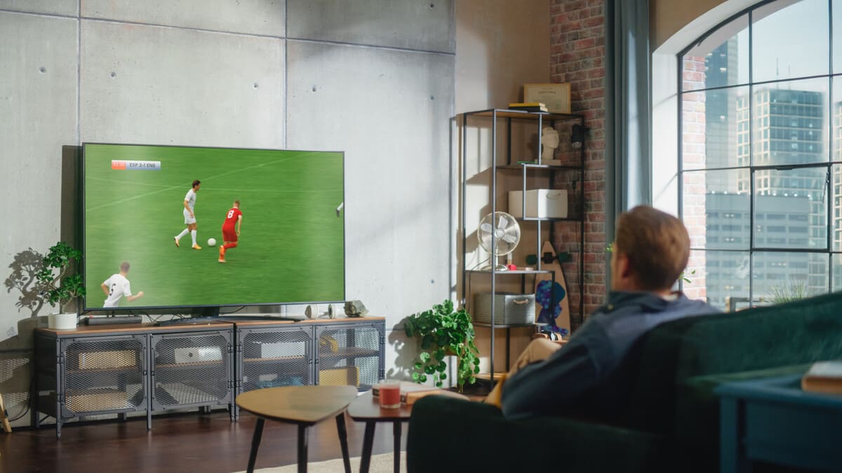 Homem assistindo a uma partida de futebol em uma TV de tela grande na sua sala de estar, um dos lugares mais confortáveis onde assistir a Copa do Mundo.
