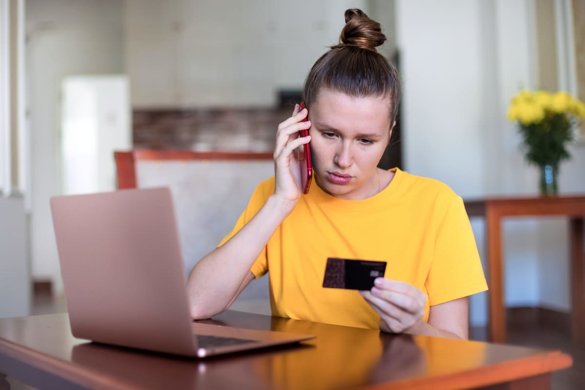 Mulher ao telefone analisando o cartão de crédito em frente ao notebook, desconfiada após comprar em site falso.