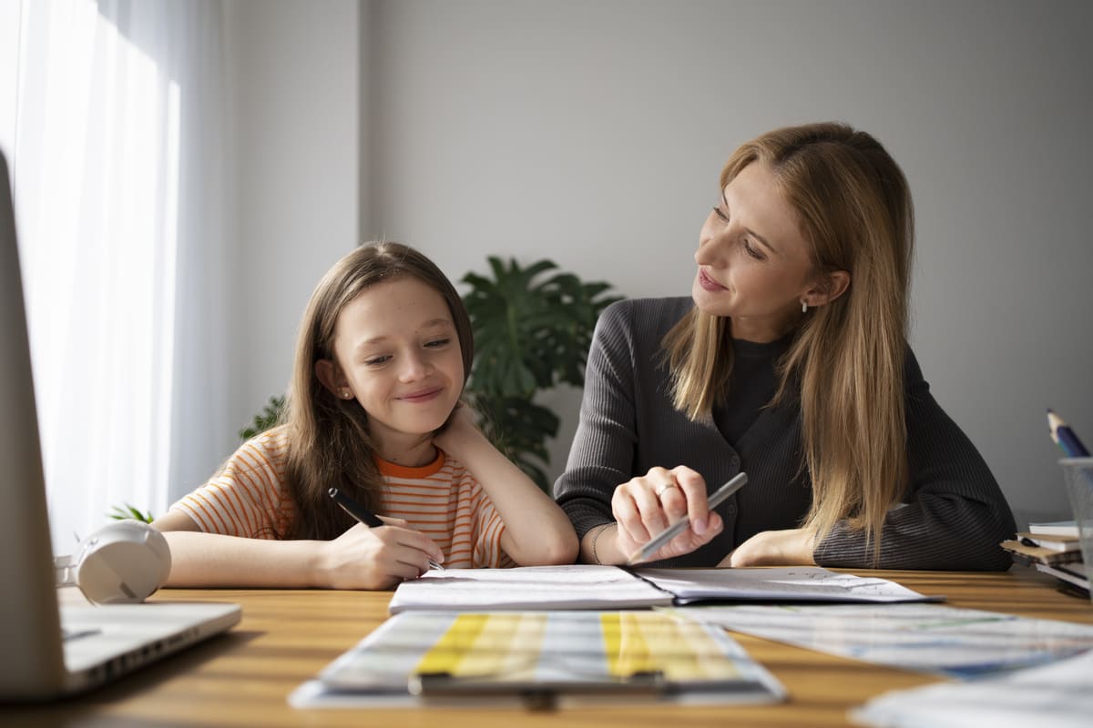 Mulher e menina sorrindo estudam com caderno e laptop sobre a mesa, focadas no aprendizado de educação financeira para jovens.