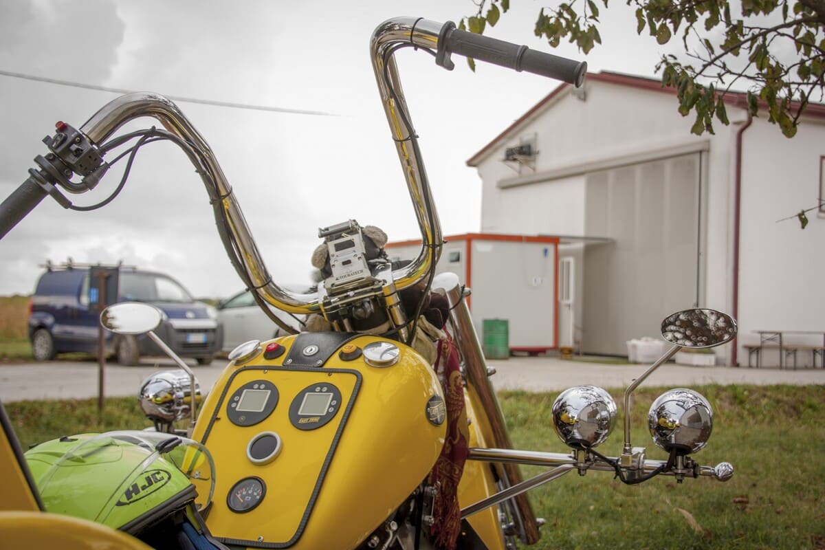 Close-up do guidão cromado de uma moto chopper amarela customizada, estacionada em uma área gramada com um galpão ao fundo.