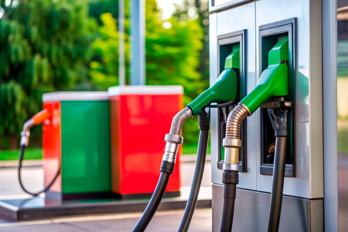 Close-up de duas pistolas de abastecimento verdes encaixadas em uma bomba de posto moderna, com outras bombas coloridas ao fundo, aludindo a dicas de como economizar combustível na estrada.