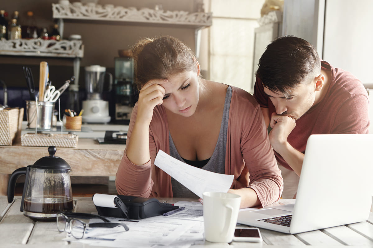 Casal jovem com a mão na cabeça analisa contas e laptop na mesa da cozinha, demonstrando frustração ao receber aviso de crédito recusado.