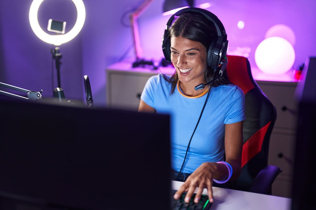 Gamer sorrindo em um ambiente com iluminação neon roxa, utilizando equipamentos para streaming essenciais, incluindo ring light e headset com microfone, durante uma transmissão no computador.