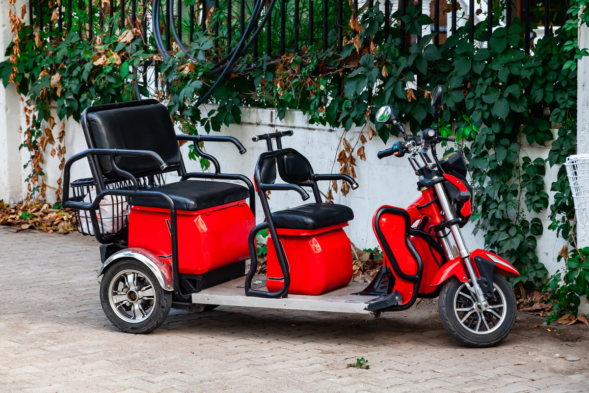 Triciclo elétrico vermelho estacionado em uma calçada pavimentada ao lado de um muro coberto por vegetação.