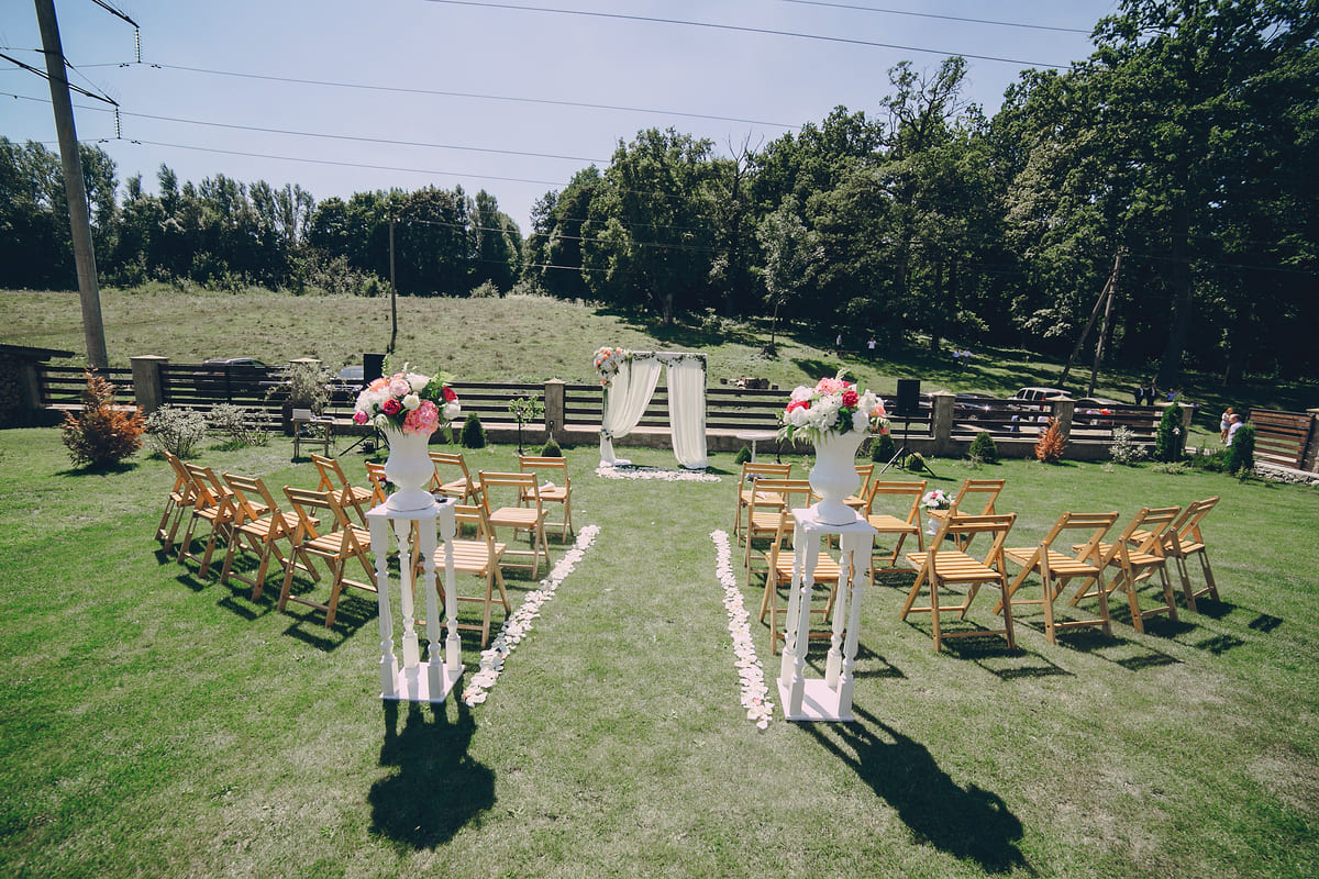 Cadeiras de madeira organizadas no gramado formando um corredor até o altar decorado com flores e tecidos, ilustrando a beleza e a preparação de um casamento ao ar livre.