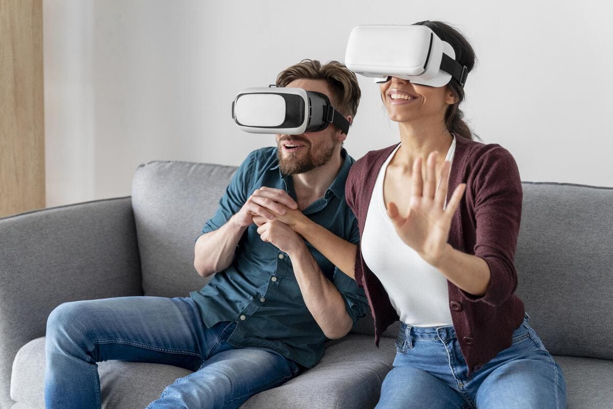 Casal sentado no sofá se diverte usando óculos VR, com a mulher estendendo a mão para interagir com o ambiente virtual.