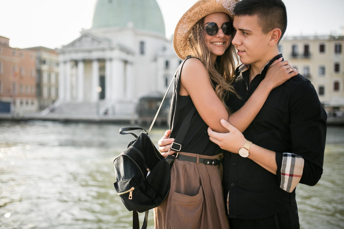 Casal abraçado e sorridente em frente a um canal com arquitetura histórica ao fundo, ilustrando o clima perfeito de uma viagem romântica para casal.