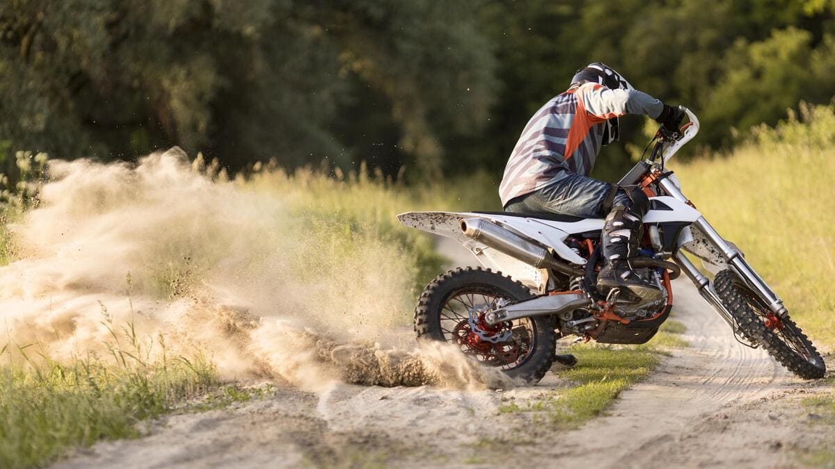 Moto enduro branca e laranja fazendo uma curva fechada e levantando poeira em uma estrada de terra.