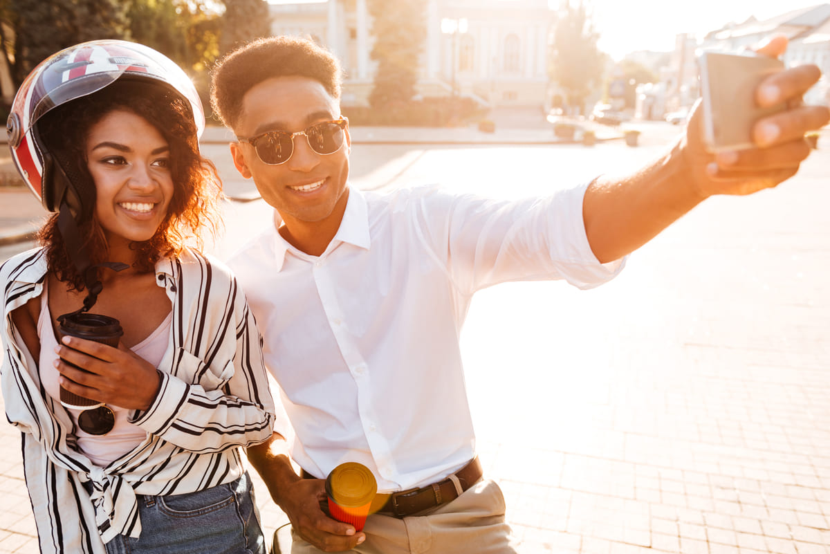 Casal jovem sorridente tirando uma selfie ao ar livre com cafés na mão, sugerindo um passeio romântico como opção de o que fazer no Dia dos Namorados.