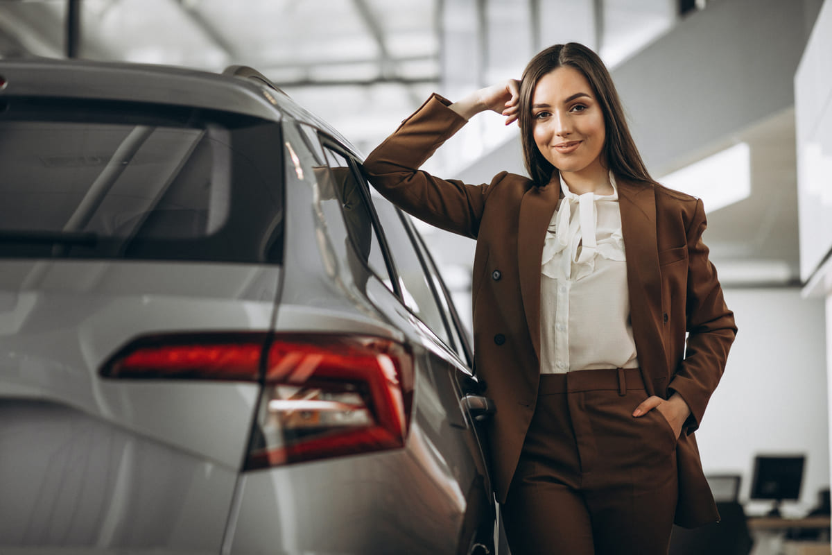 Mulher elegante com terno marrom encostada em um veículo novo dentro de uma concessionária, refletindo sobre qual a melhor forma de comprar um carro.