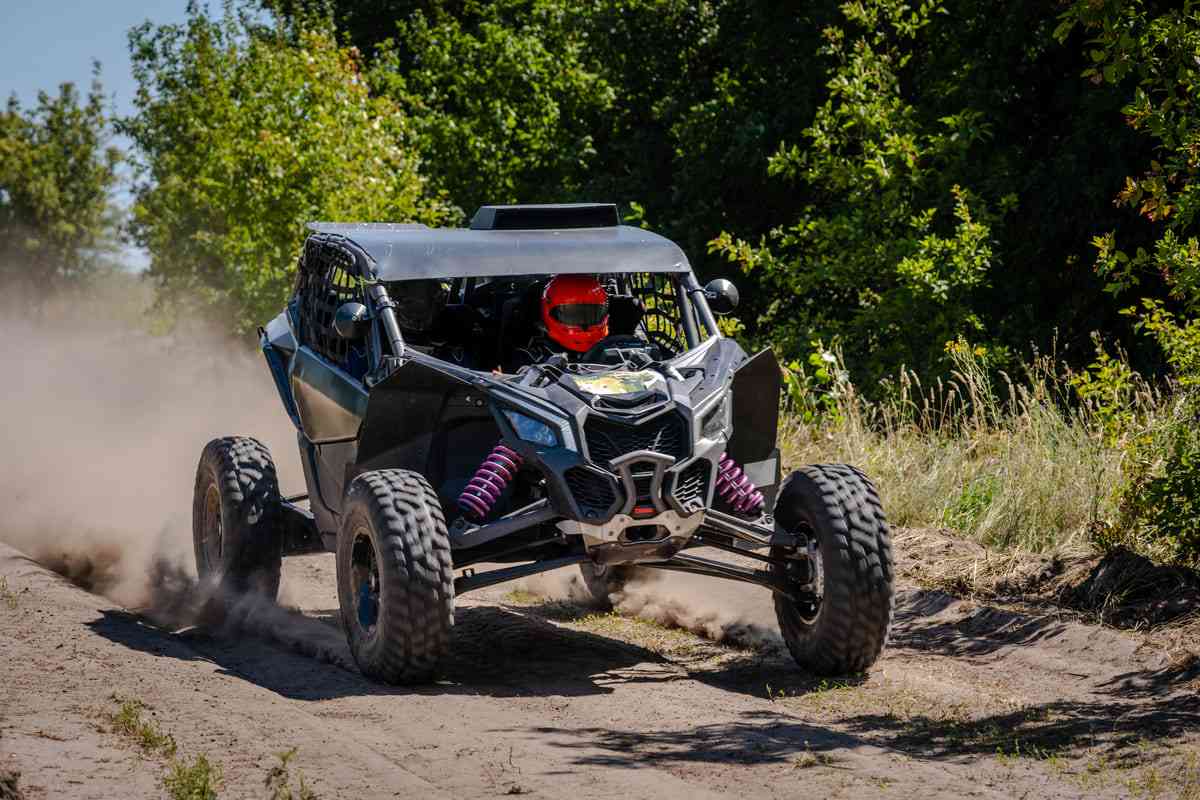 Piloto com capacete vermelho corre com veículo utv em trilha levantando poeira.