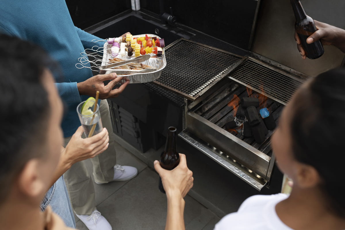 Pessoas reunidas com bebidas e espetinhos ao redor de uma churrasqueira em apartamento.